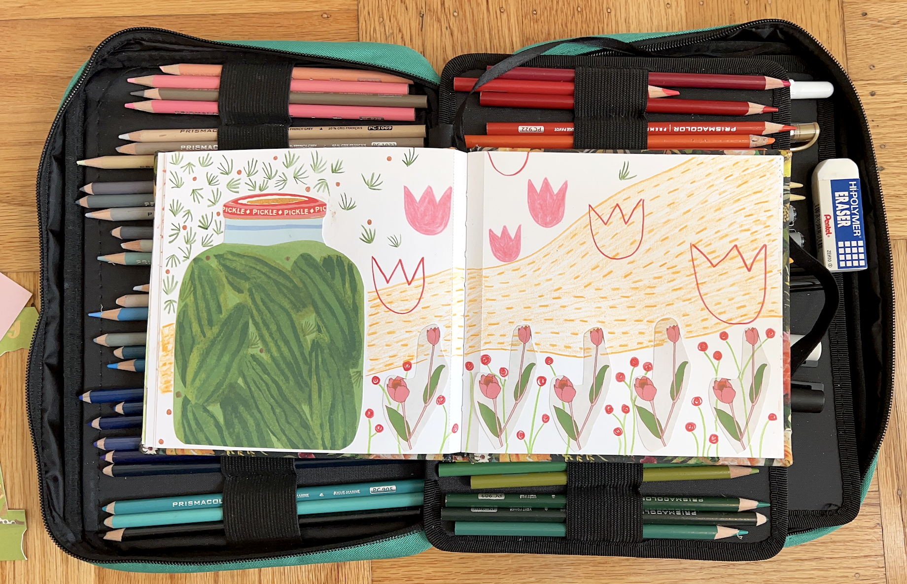 image of an open sketchbook with collage and pencil of florals and pickles on top of a travel-friendly pencil case on wood floor
